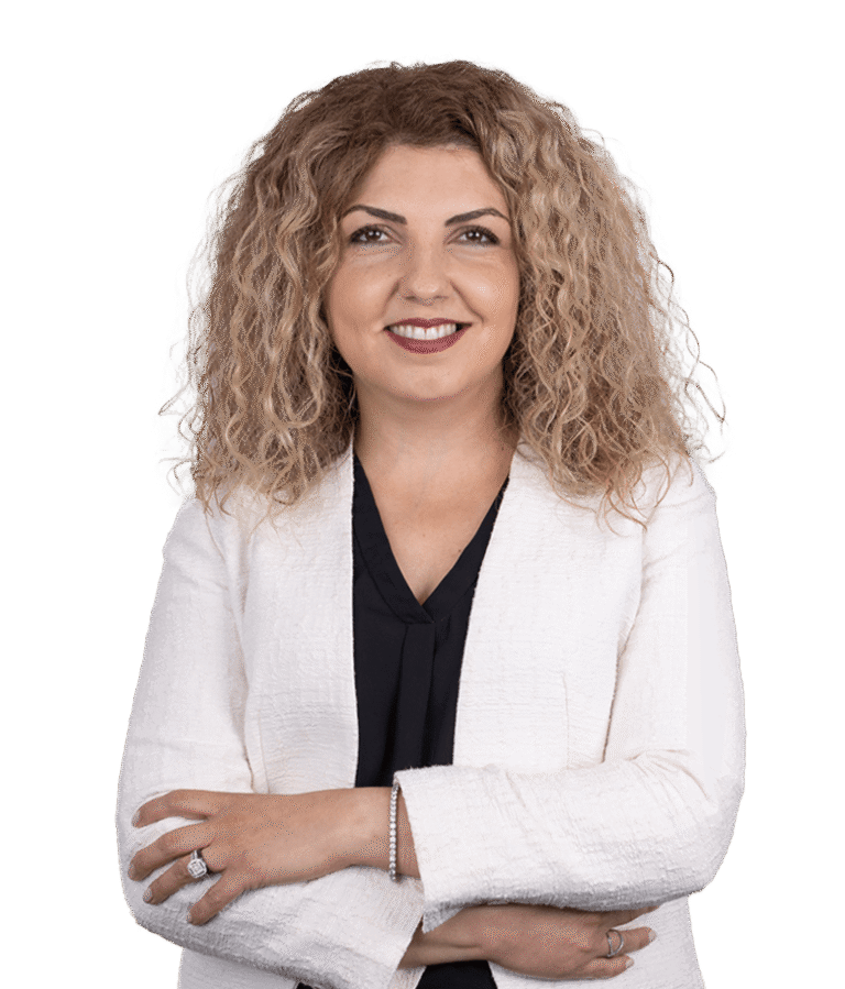 A woman with curly, shoulder-length blonde hair smiles at the camera. She is wearing a white blazer over a black top and has her arms crossed. The background is plain white.