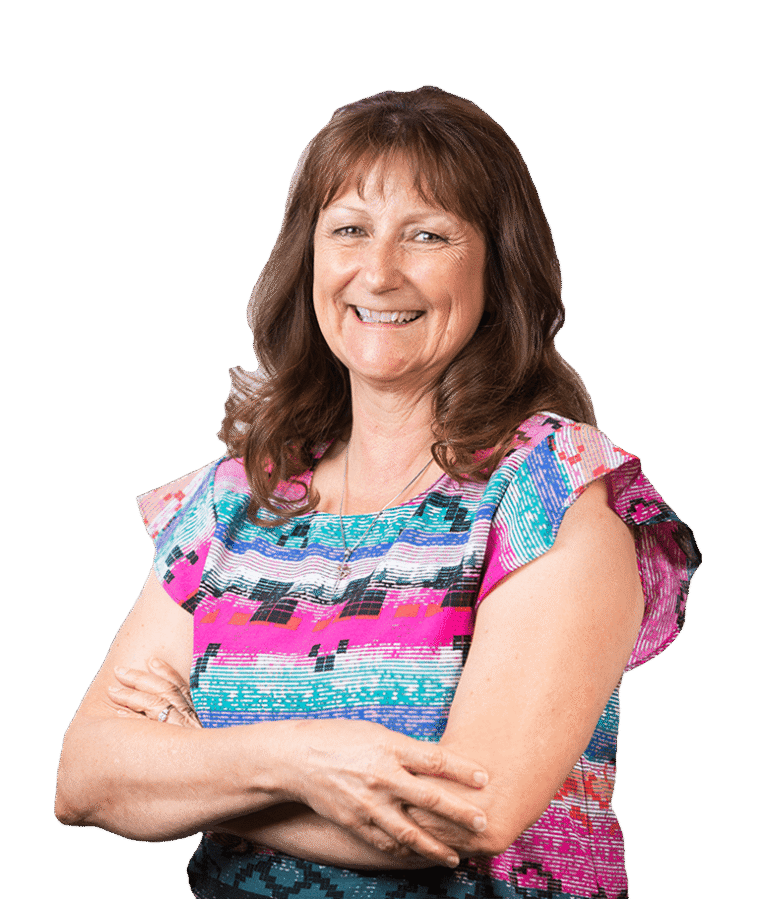 A smiling woman with wavy brown hair stands with her arms crossed, wearing a colorful, patterned blouse in shades of pink, blue, and white, against a plain white background.