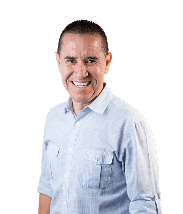 A man with short dark hair, wearing a light blue button-up shirt, smiles warmly at the camera against a plain white background.