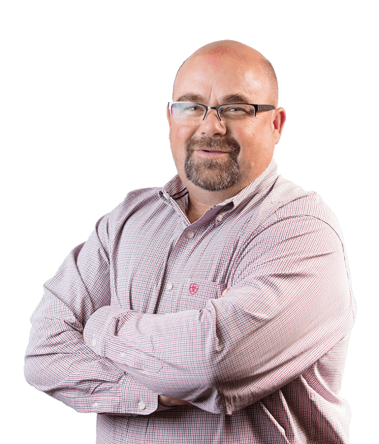 A man with a trimmed beard and glasses, wearing a light plaid button-up shirt, stands with arms crossed and smiles confidently against a plain white background.