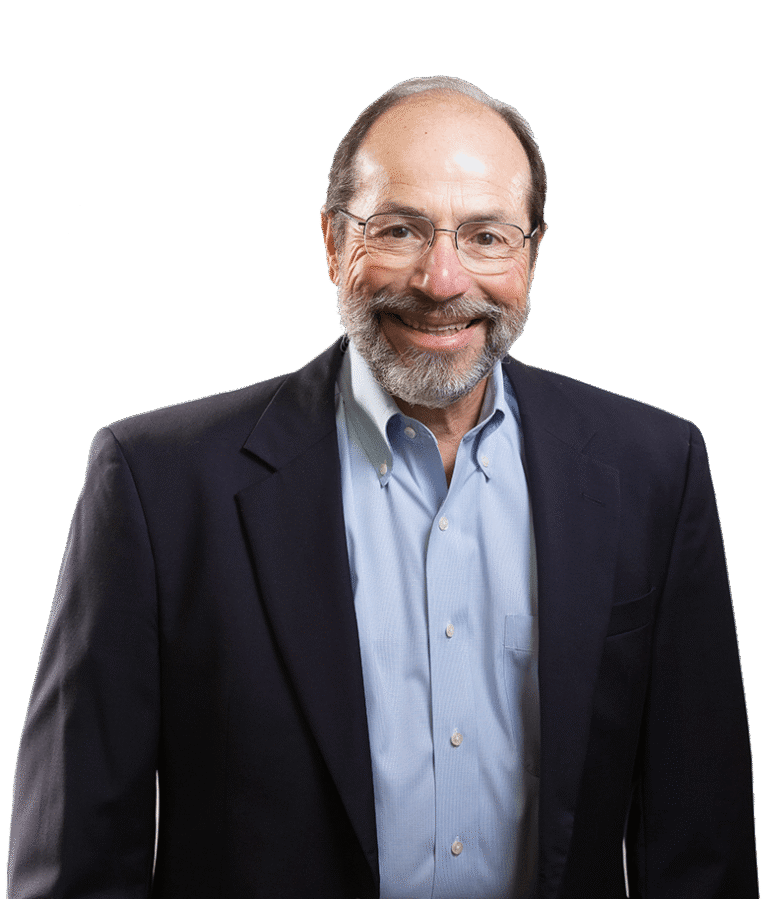 A smiling older man with glasses, a beard, and mustache, wearing a dark suit jacket over a light blue button-up shirt, stands against a plain white background.