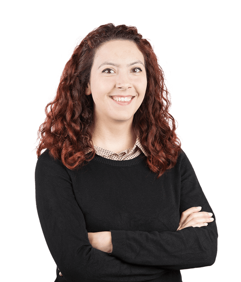A woman with curly red hair smiles with her arms crossed, wearing a black sweater over a collared shirt, standing against a plain white background.