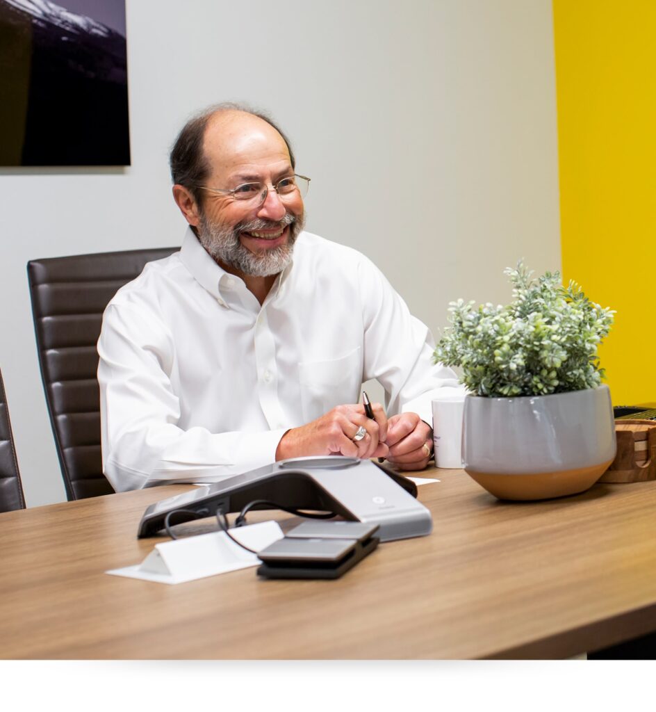 A smiling man with glasses and a beard sits at a modern desk, writing with a pen. The desk has a plant, a phone, and office supplies. The background features a white and yellow wall.