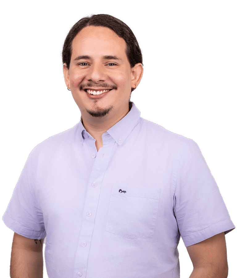 A man with dark hair and a goatee, wearing a light purple button-up shirt, smiles at the camera against a plain white background.