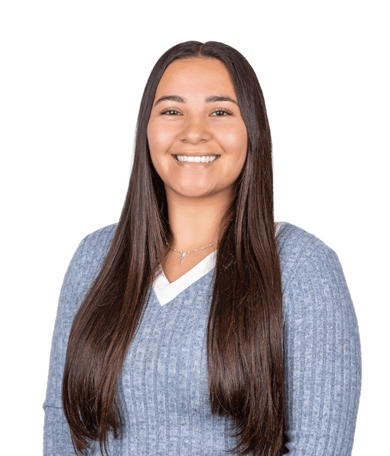 A woman with long straight brown hair, wearing a light blue ribbed sweater with a white neckline and a silver necklace, smiles at the camera against a plain white background.