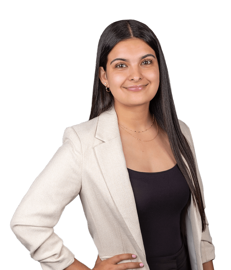 A woman with long dark hair, wearing a light beige blazer over a black top, standing with one hand on her hip and smiling against a plain white background.