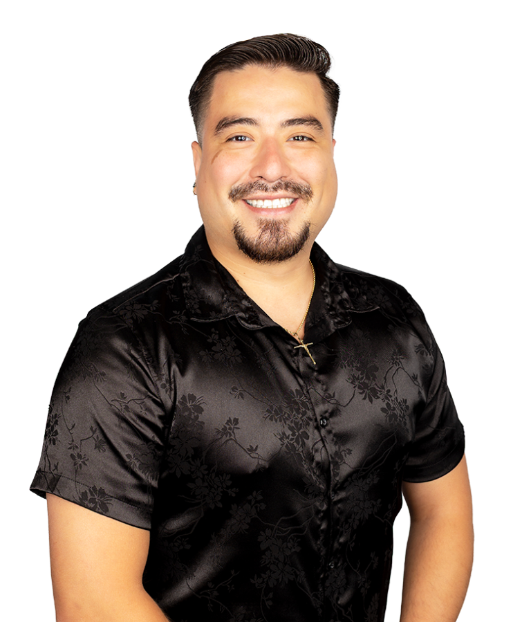 Smiling man with short dark hair and a goatee wearing a black, short-sleeved, floral-patterned shirt and a gold necklace, standing against a plain white background.