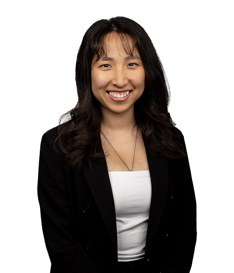 A woman with long dark hair smiles at the camera. She is wearing a black blazer over a white top and a necklace, standing against a plain white background.