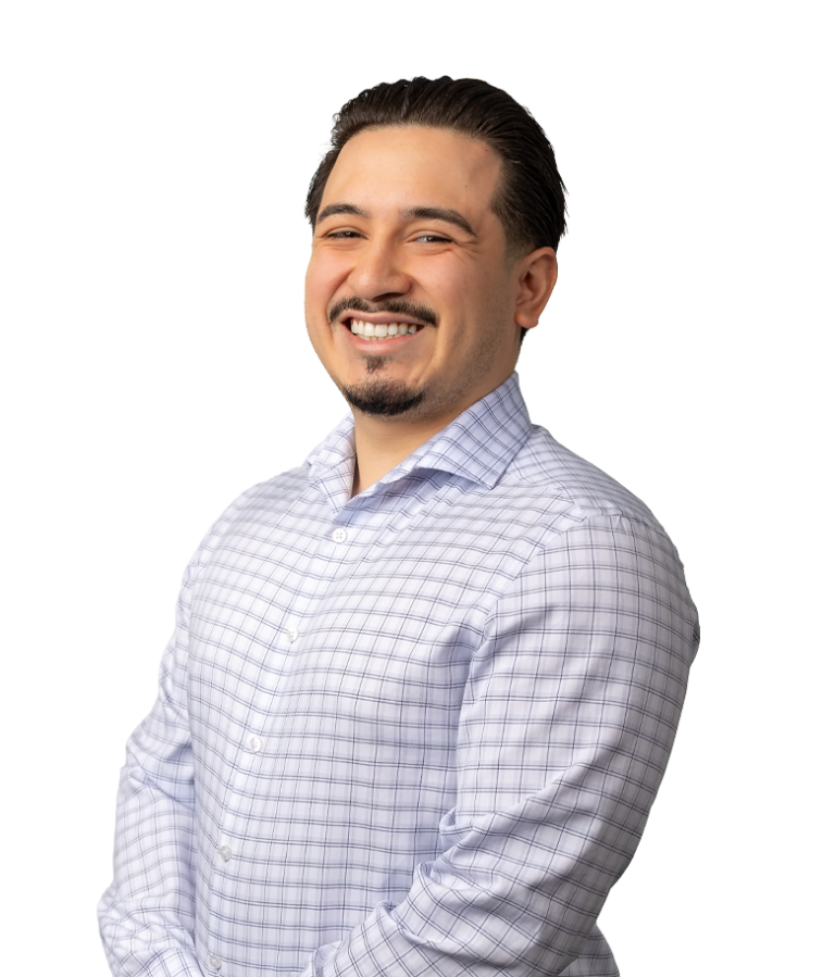 A smiling man with slicked-back dark hair and a goatee, wearing a light-colored, checkered dress shirt, stands against a plain white background.
