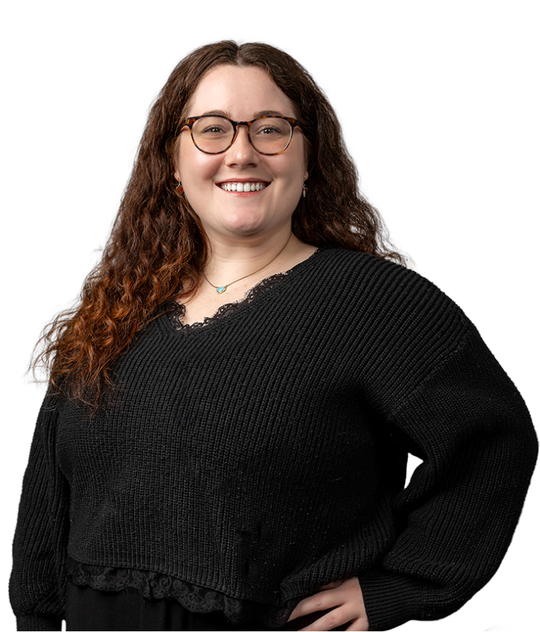 A smiling woman with long, wavy brown hair and glasses, wearing a black knit sweater and a necklace, stands against a plain white background with one hand on her hip.