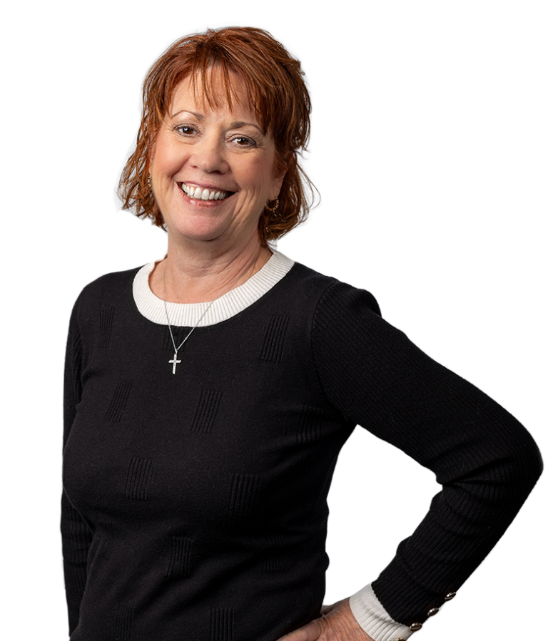 Smiling woman with short reddish hair wearing a black sweater with white collar and cuffs, and a cross necklace, standing with one hand on her hip against a plain white background.