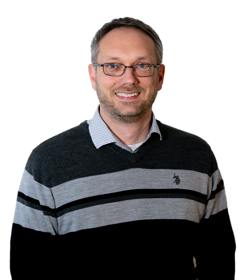 A smiling man with short brown hair, glasses, and a beard is wearing a black and gray striped sweater over a collared shirt, standing against a plain white background.